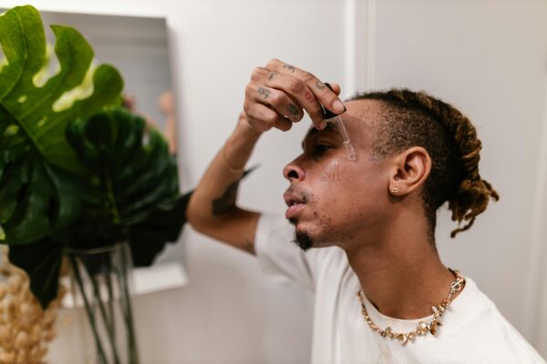 A young man with tattoos and dreadlocks applies skincare serum to his face indoors.