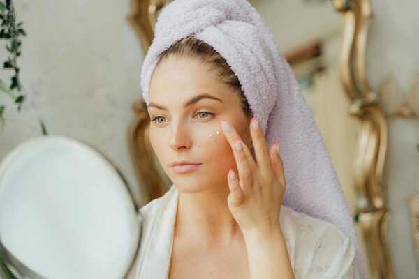 Woman with head towel applying moisturizer in front of mirror indoors.