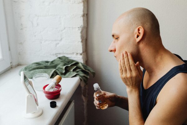 Bald man applying toner to face beside mirror with skincare products in bathroom.